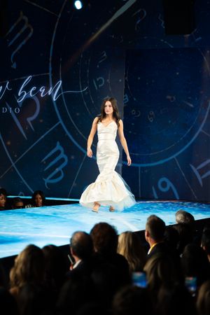 Model walks runway in elegant white mermaid gown at a bridal fashion show.
