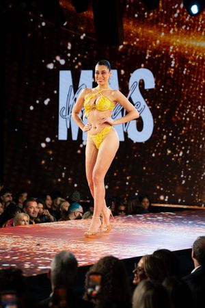 Model walks runway in yellow bikini at fashion show with golden backdrop and audience.