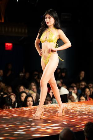 Model showcases yellow bikini set on illuminated fashion runway before seated audience.
