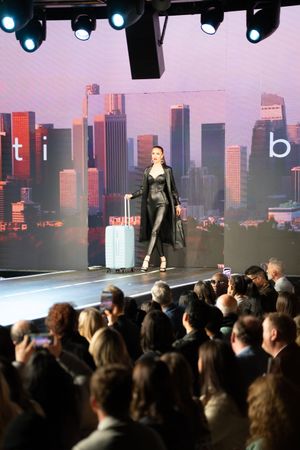 Model in black leather outfit walks runway with white luggage, city skyline backdrop, fashion show audience.
