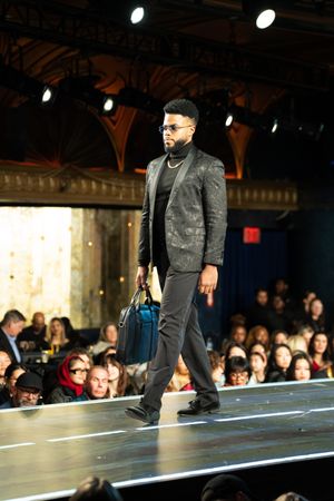 Male model carries blue bag down runway wearing dark patterned blazer and slim trousers.