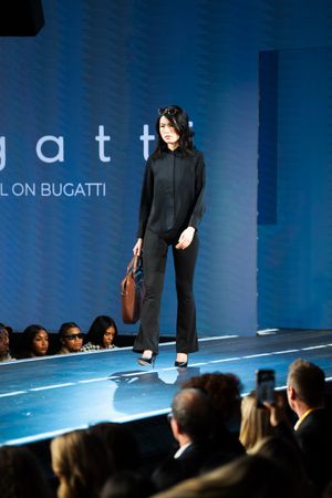 Model walks Bugatti fashion show runway in all-black outfit carrying brown leather tote bag.