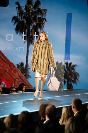 Female model walks runway in tan mink fur coat carrying gray handbag at Bucatti fashion show.