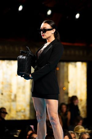 Female model walks runway in black blazer dress holding black handbag wearing sunglasses.