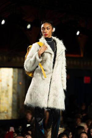 Female model poses on runway wearing white faux fur coat with yellow backpack and black leather pants.