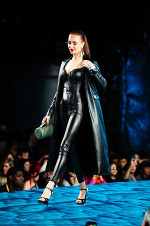 Model showcases black leather catsuit and long coat ensemble on blue-lit runway at fashion show.