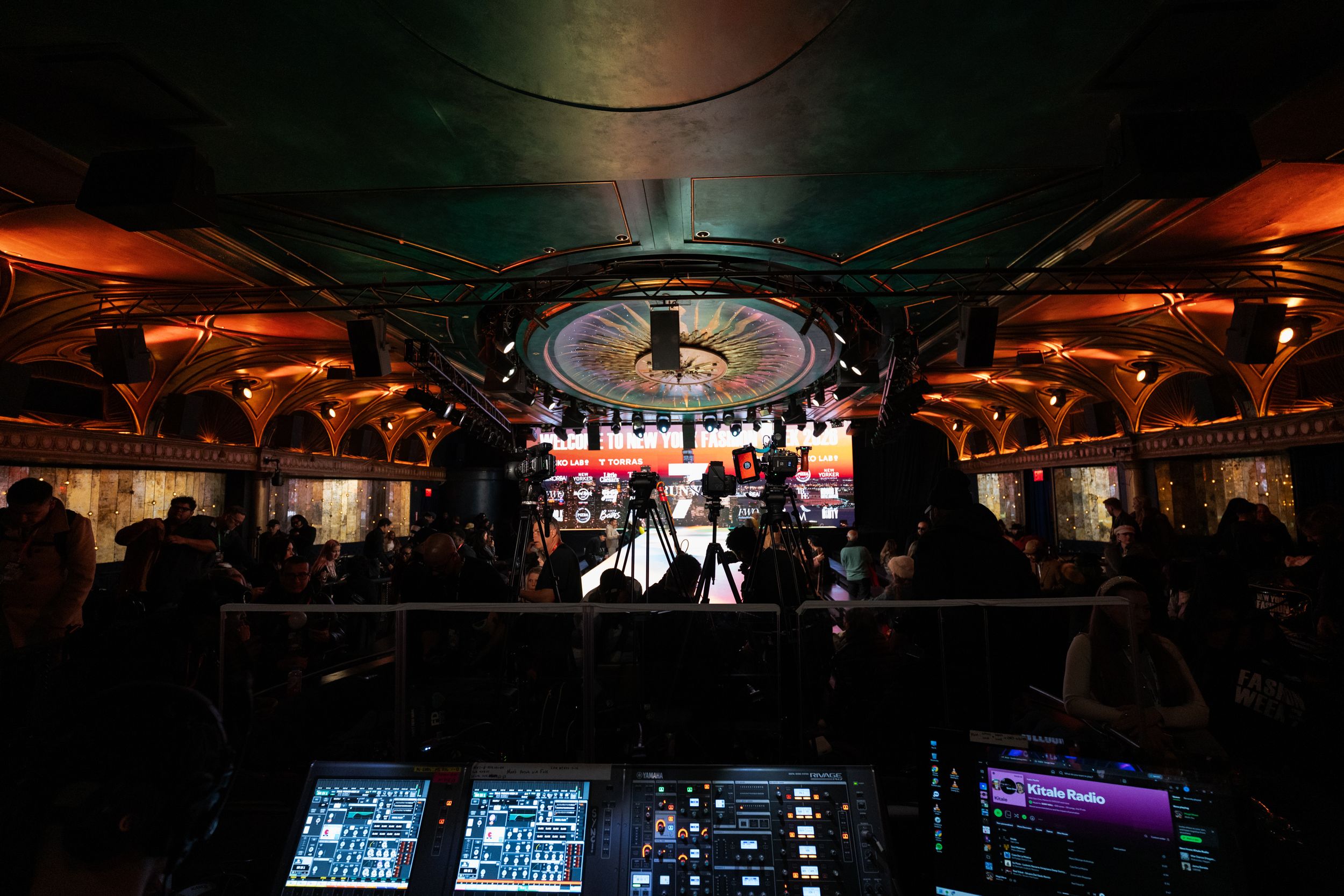 DJ booth overlooking crowded nightclub with ornate ceiling and colorful stage lighting.