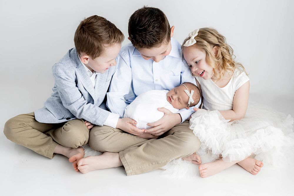 Three siblings sit together on white backdrop, two boys in blue shirts lovingly cradling a swaddled newborn baby.