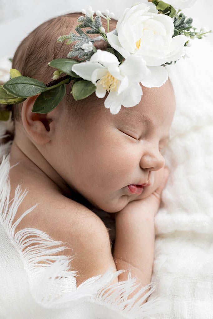 Sleeping newborn baby wearing a white floral crown, resting peacefully on a soft white blanket.