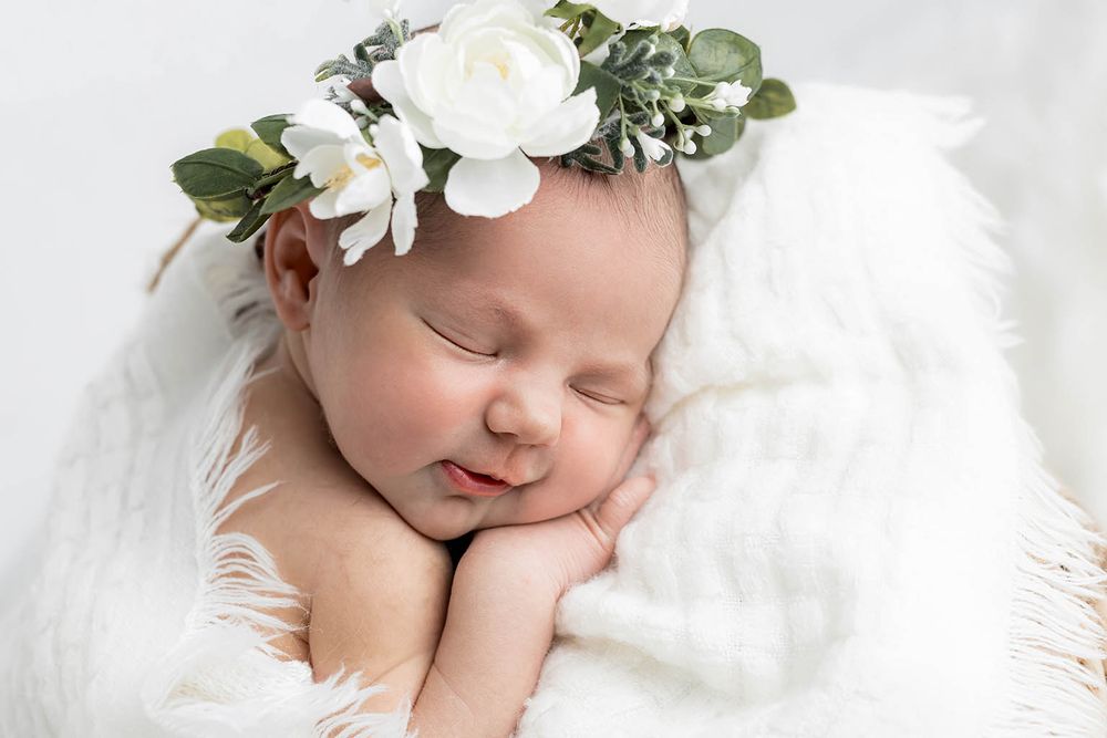 Sleeping newborn baby girl wearing a delicate white floral crown, wrapped in soft white fabric.