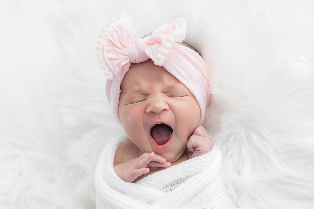 Newborn baby girl swaddled in white wrap with pink bow headband, yawning cutely on fluffy white backdrop.