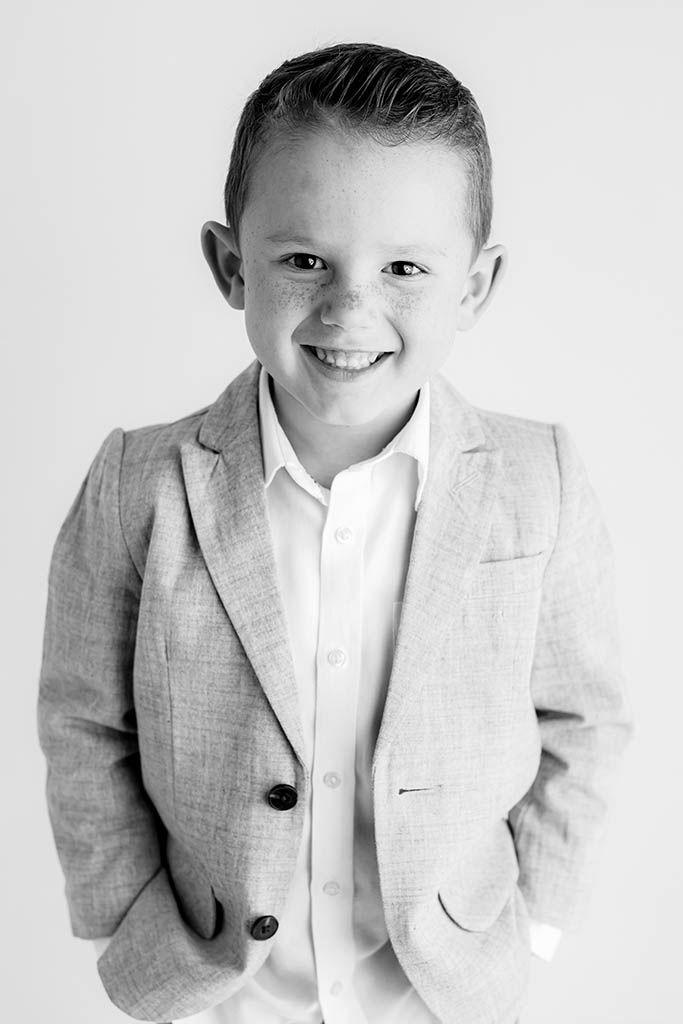 A cheerful young boy in a gray blazer and white shirt smiles confidently in a black and white portrait.