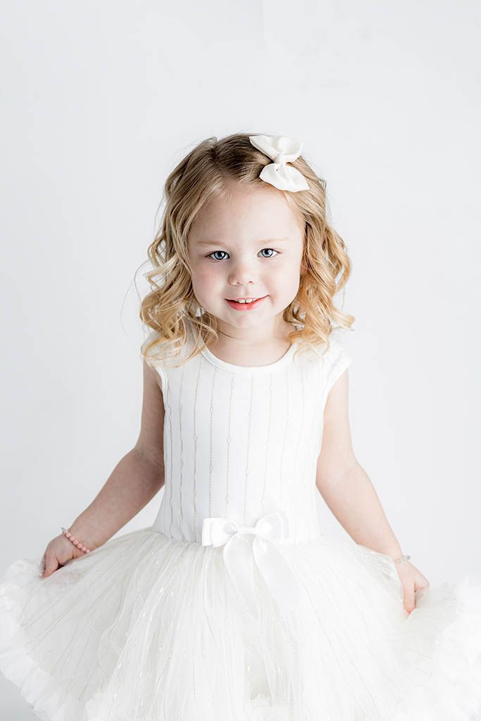 Smiling little girl in white tulle dress and bow hair clip poses against a light background.