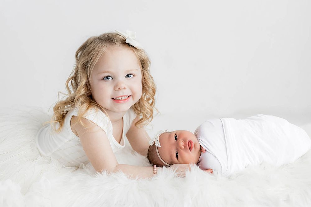 Young girl in white dress smiles beside swaddled newborn baby on fluffy white blanket in bright studio photo.