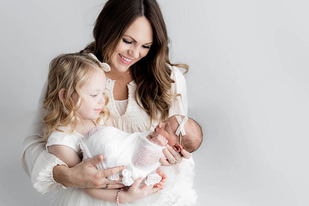 A smiling mother in white holds her newborn baby and young blonde daughter in matching white dresses.
