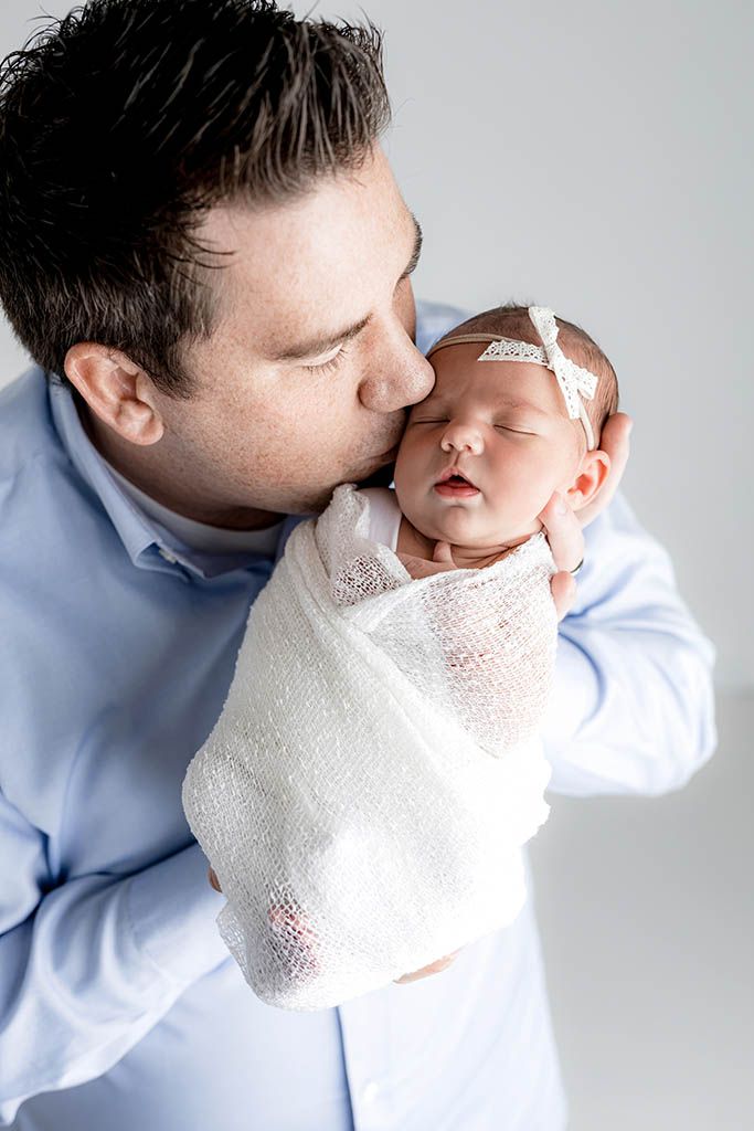 A father in a blue shirt tenderly kisses his swaddled newborn baby wrapped in white, against a light background.