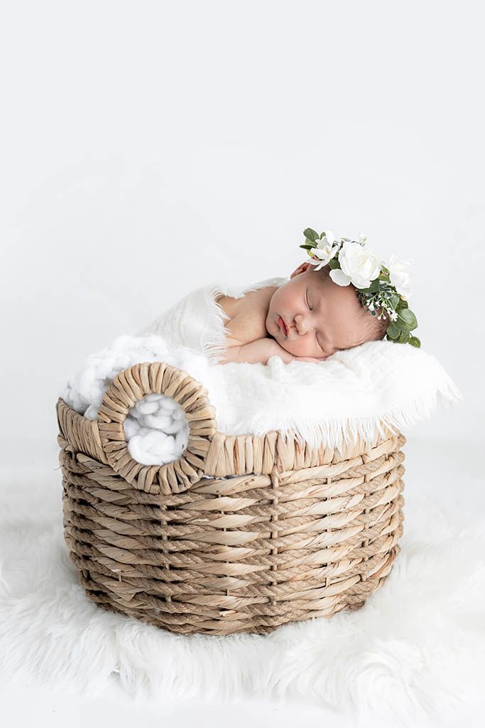 Sleeping newborn baby wearing a white floral crown resting in a wicker basket lined with soft white fur blanket.