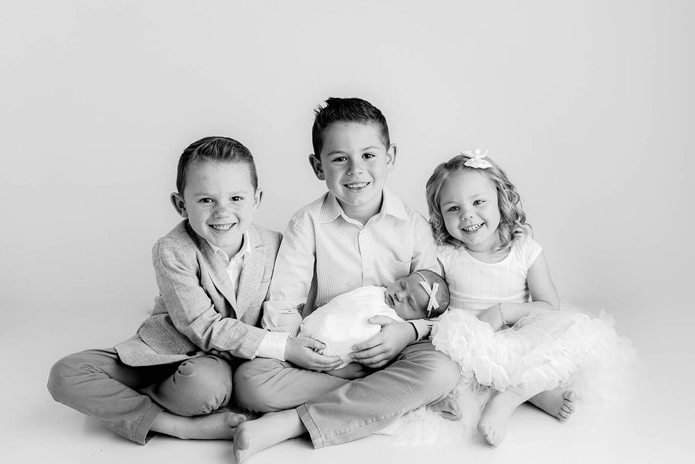 Three siblings smile together in black and white photo, oldest boy holding sleeping newborn baby.