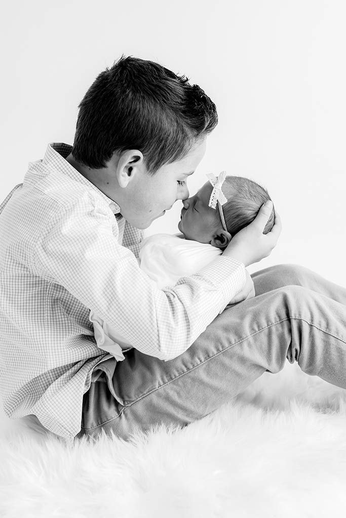 Young boy tenderly holds and gazes at newborn baby sister in black and white portrait.