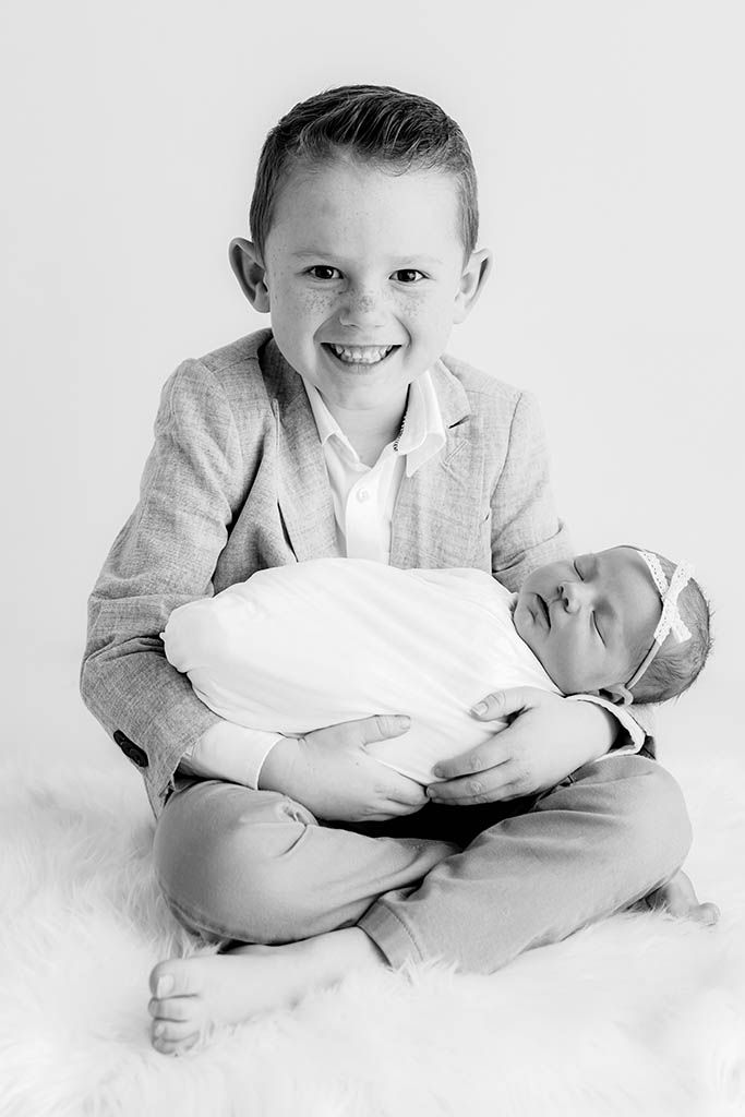 Smiling boy in blazer holds sleeping newborn baby sister in classic black and white photo by Menifee family and newborn photographer.