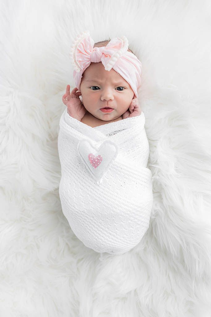 Newborn baby girl swaddled in white wrap with heart detail, wearing pink bow headband on fluffy white rug.