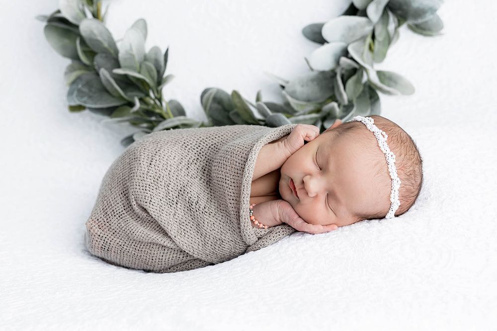 Sleeping newborn baby swaddled in gray knit wrap posed on white blanket with green eucalyptus wreath.