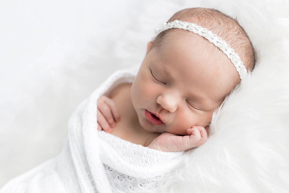 Peaceful newborn baby sleeping in white swaddle wrap wearing delicate lace headband on white background.