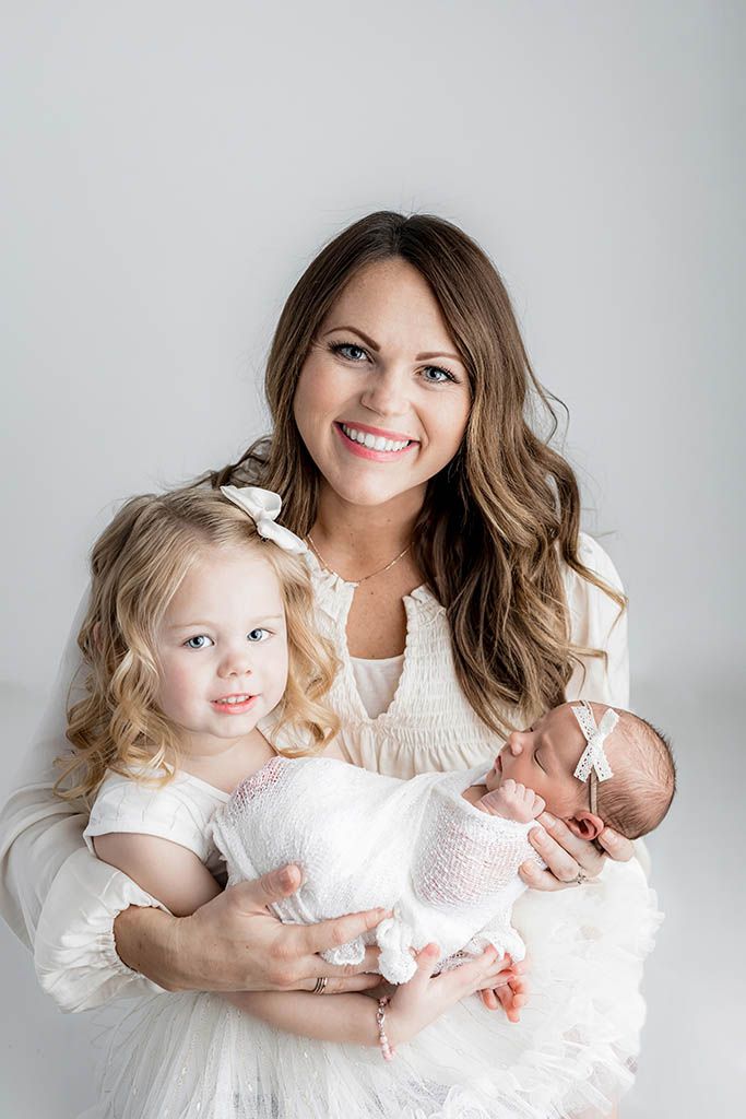 Smiling mother holds toddler girl and swaddled newborn baby in studio portrait by Menifee family and newborn photographer.