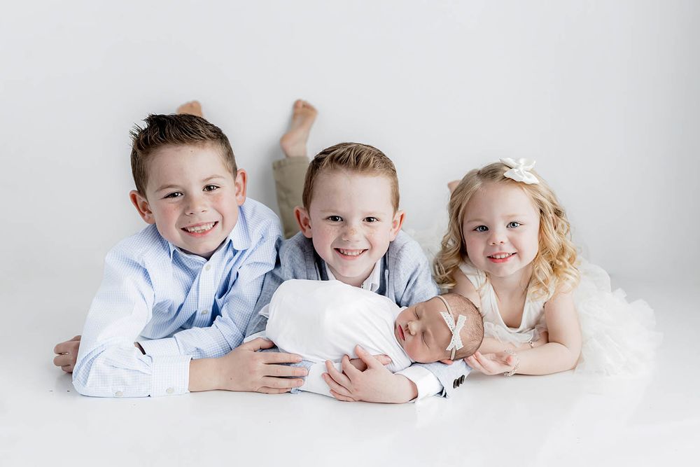 Three smiling siblings in blue and white outfits lying down together with swaddled newborn baby by Menifee family and newborn photographer.
