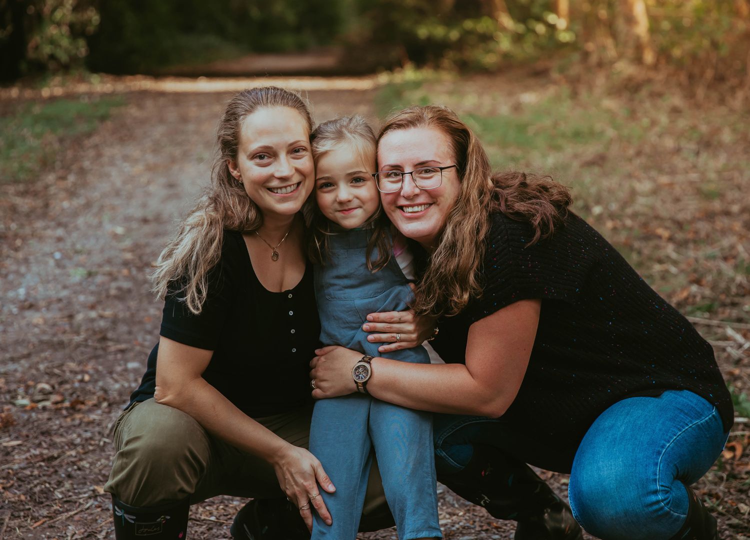 Two women and a young girl smile together on a wooded dirt path in a warm outdoor family portrait. During a family photoshoot in Orpington