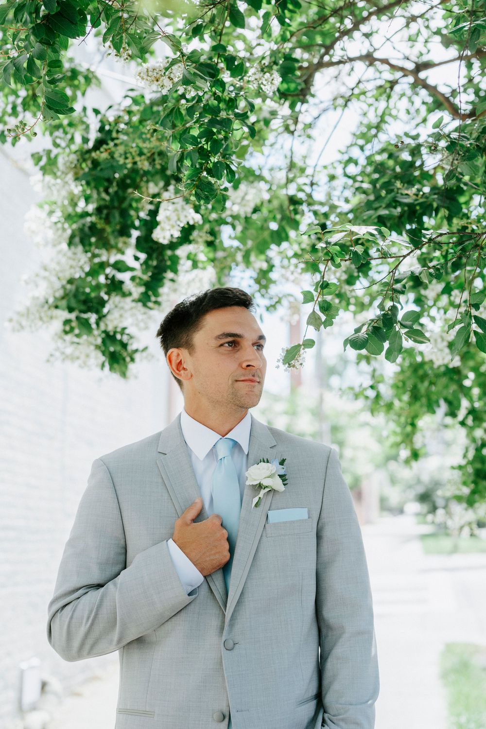 Groom in light gray suit with boutonniere standing under flowering tree branches on wedding day.