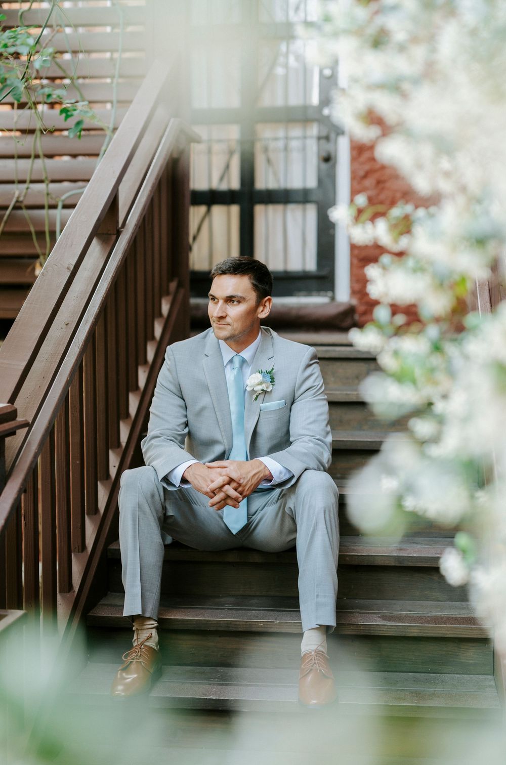 Groom in light gray suit with boutonniere sits on wooden steps outdoors, looking thoughtfully to the side.