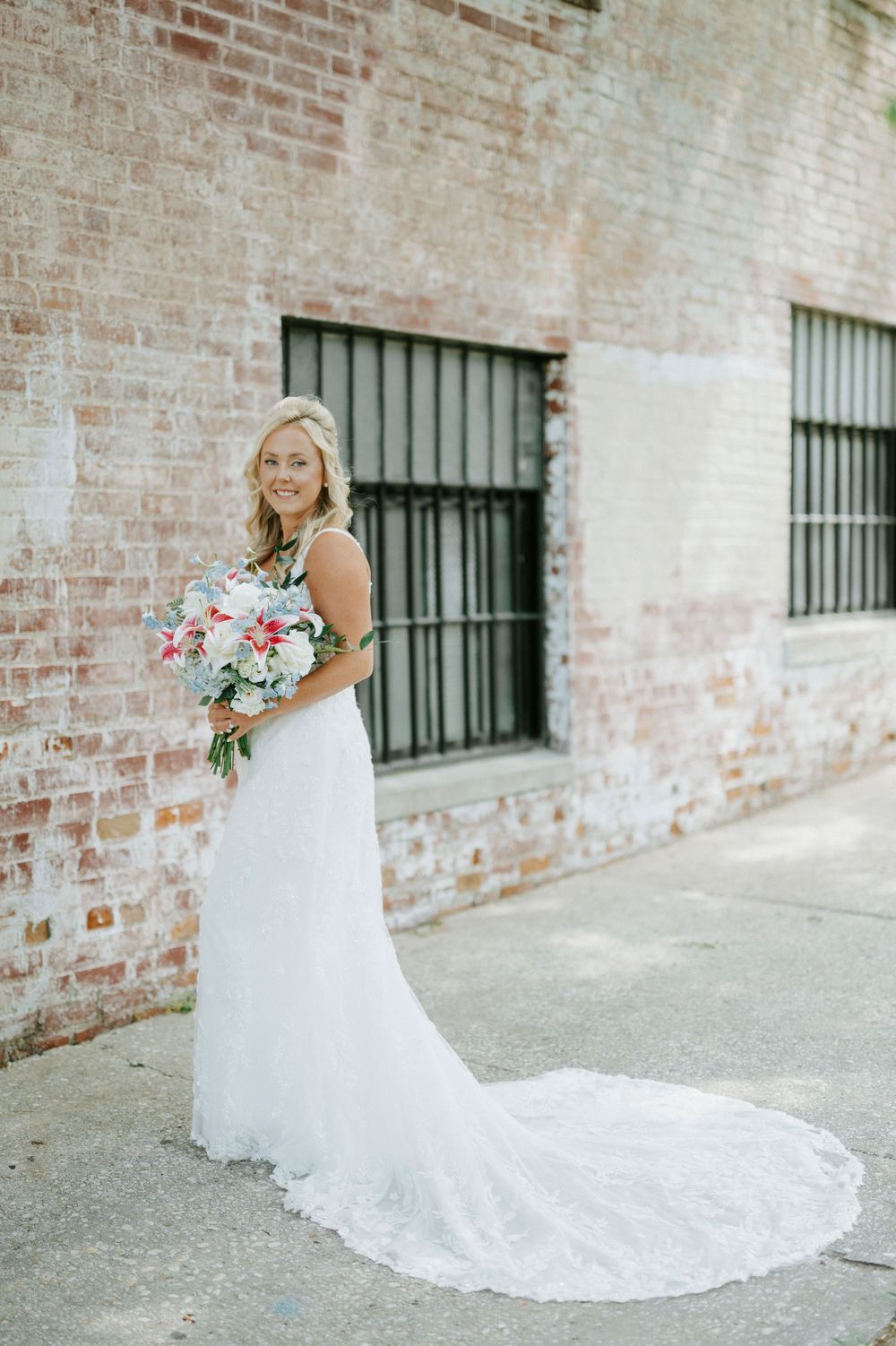 Bride in white lace gown with train holds colorful bouquet in front of rustic brick wall.
