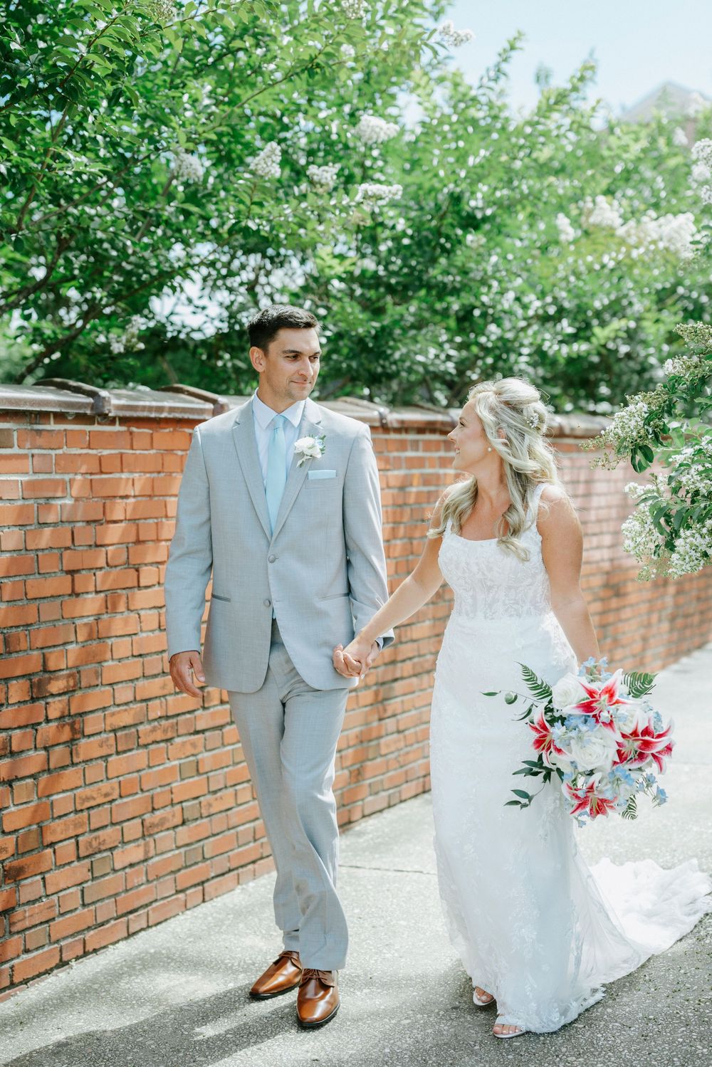 Bride and groom walking hand in hand beside brick wall with lush greenery and bridal bouquet.