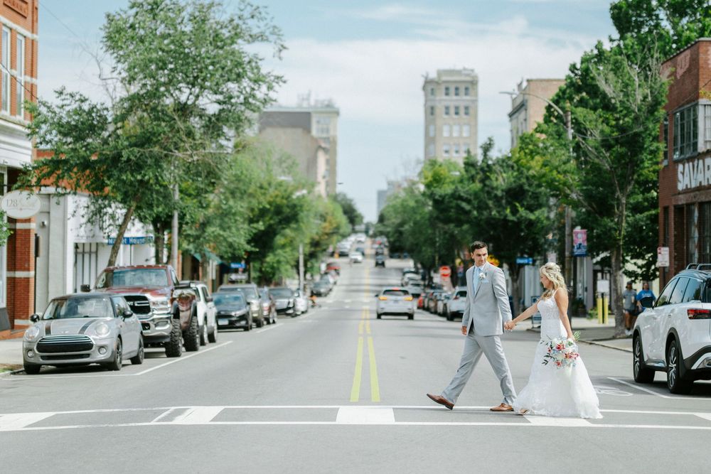 Bride and groom crossing a sunny downtown street in wedding attire, with historic buildings and parked cars lining the road.