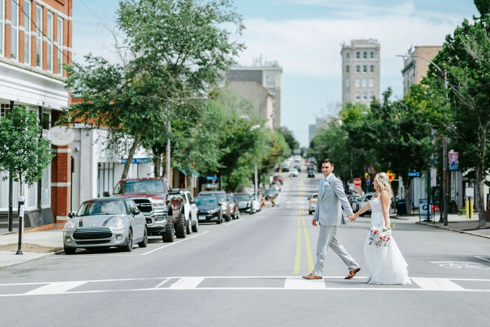 Bride and groom crossing a city street in wedding attire, with urban buildings and parked cars in the background.
