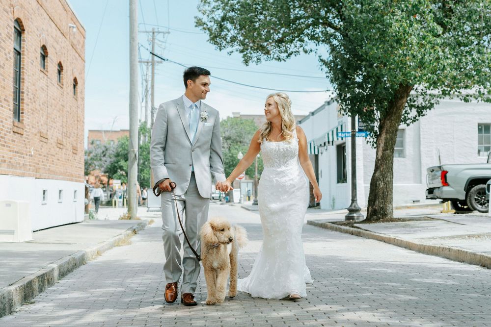 Bride and groom walking a golden doodle dog on a sunny small-town street, holding hands in wedding attire.