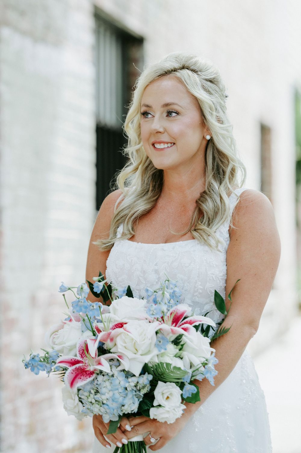 Smiling bride with blonde wavy hair holding a bouquet of pink, white, and blue flowers in a white wedding dress.