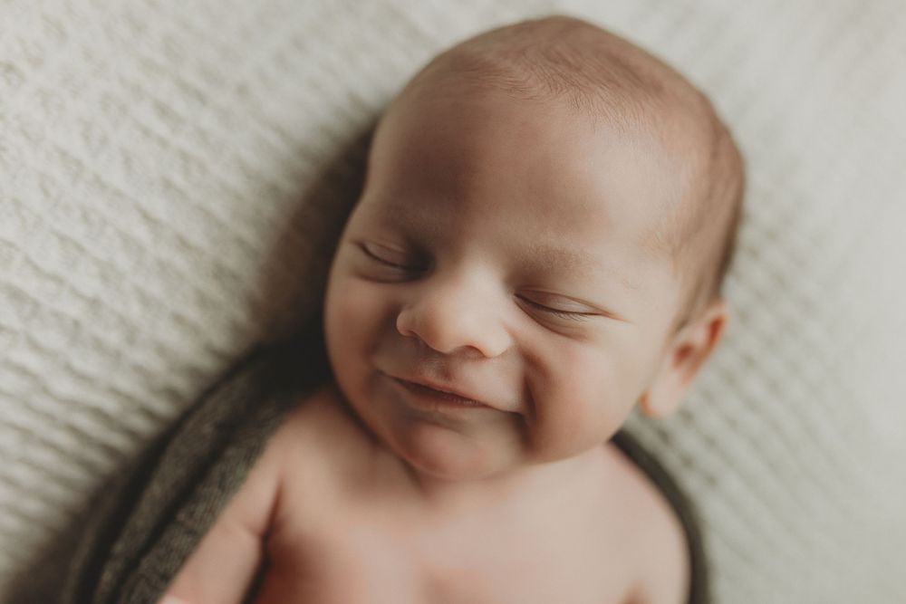 Smiling newborn baby wrapped in a soft gray blanket, peacefully sleeping on a white textured backdrop.