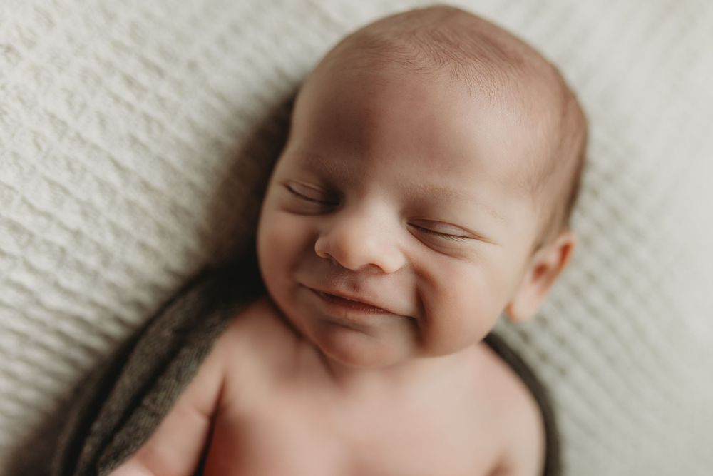 Peaceful newborn baby smiling while sleeping, wrapped in a soft gray blanket on a white textured background.