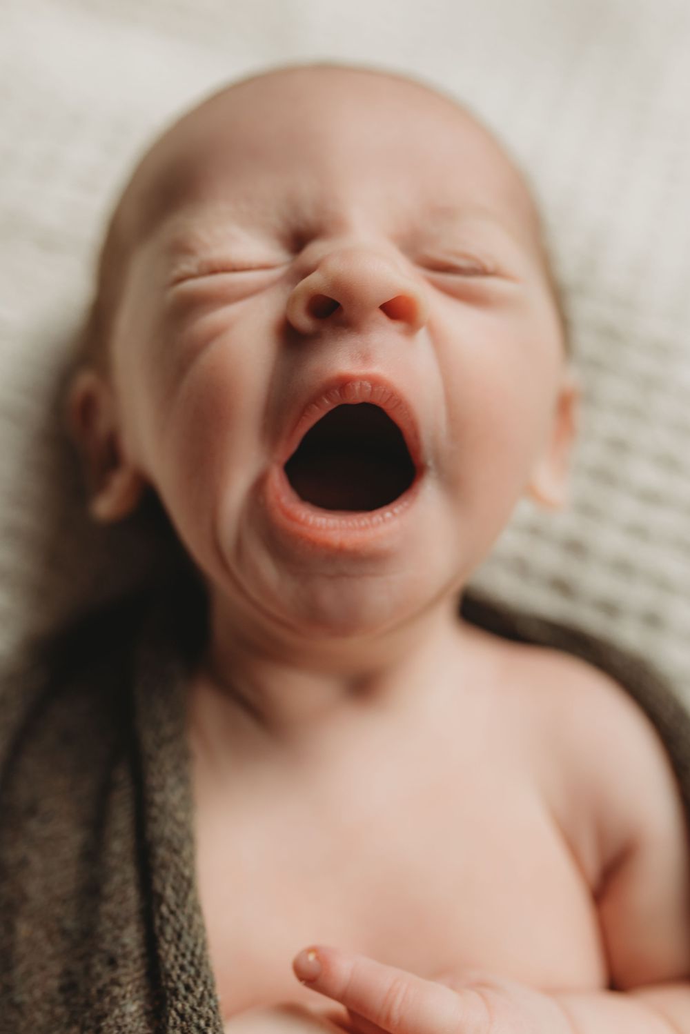 Close-up of a newborn baby yawning with eyes closed, wrapped in a soft dark blanket on a white surface.