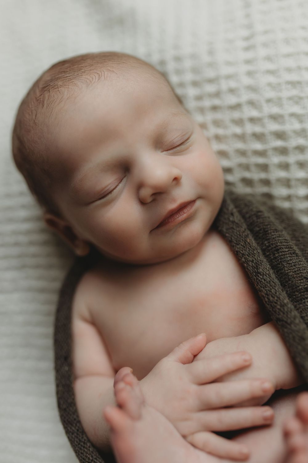 Close-up of smiling sleeping newborn wrapped in dark green swaddle on soft white blanket.
