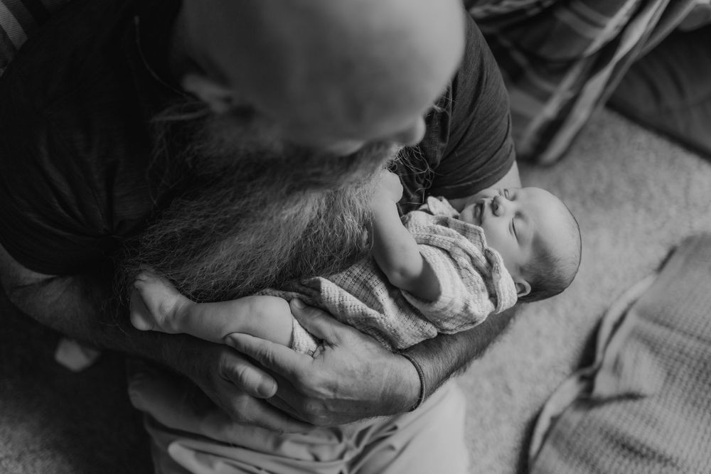 Bald father with beard kissing newborn baby held in his arms in a tender black and white portrait.