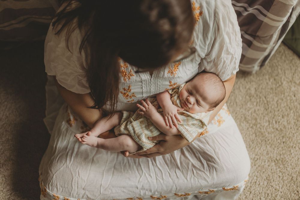 A mother cradles her sleeping newborn baby in her arms while sitting on a bed in soft natural light.