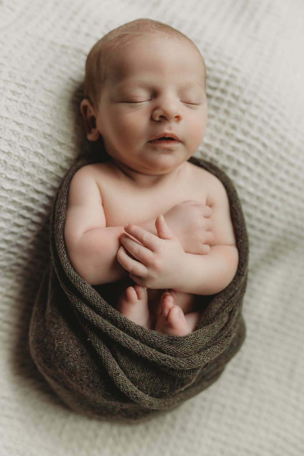 Peaceful newborn baby swaddled in olive green wrap sleeping on white textured blanket.