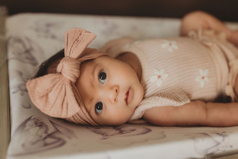 Adorable baby girl with blue eyes wearing a pink floral romper and bow headband lying on a soft blanket.