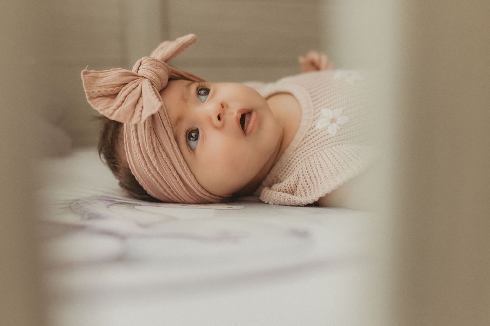 Adorable baby girl in pink bow headband lying in a white crib, looking up with big blue eyes.