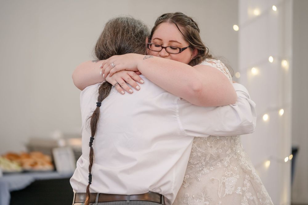 Bride in lace wedding gown hugging a guest with braided hair at a warmly lit wedding reception.