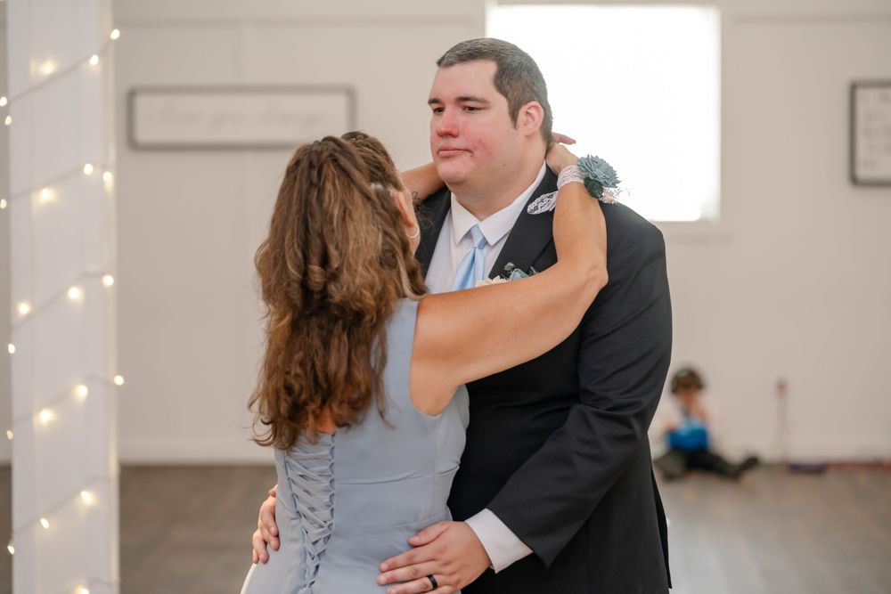 Groom in black suit shares emotional mother-son dance at elegant wedding reception with string lights.