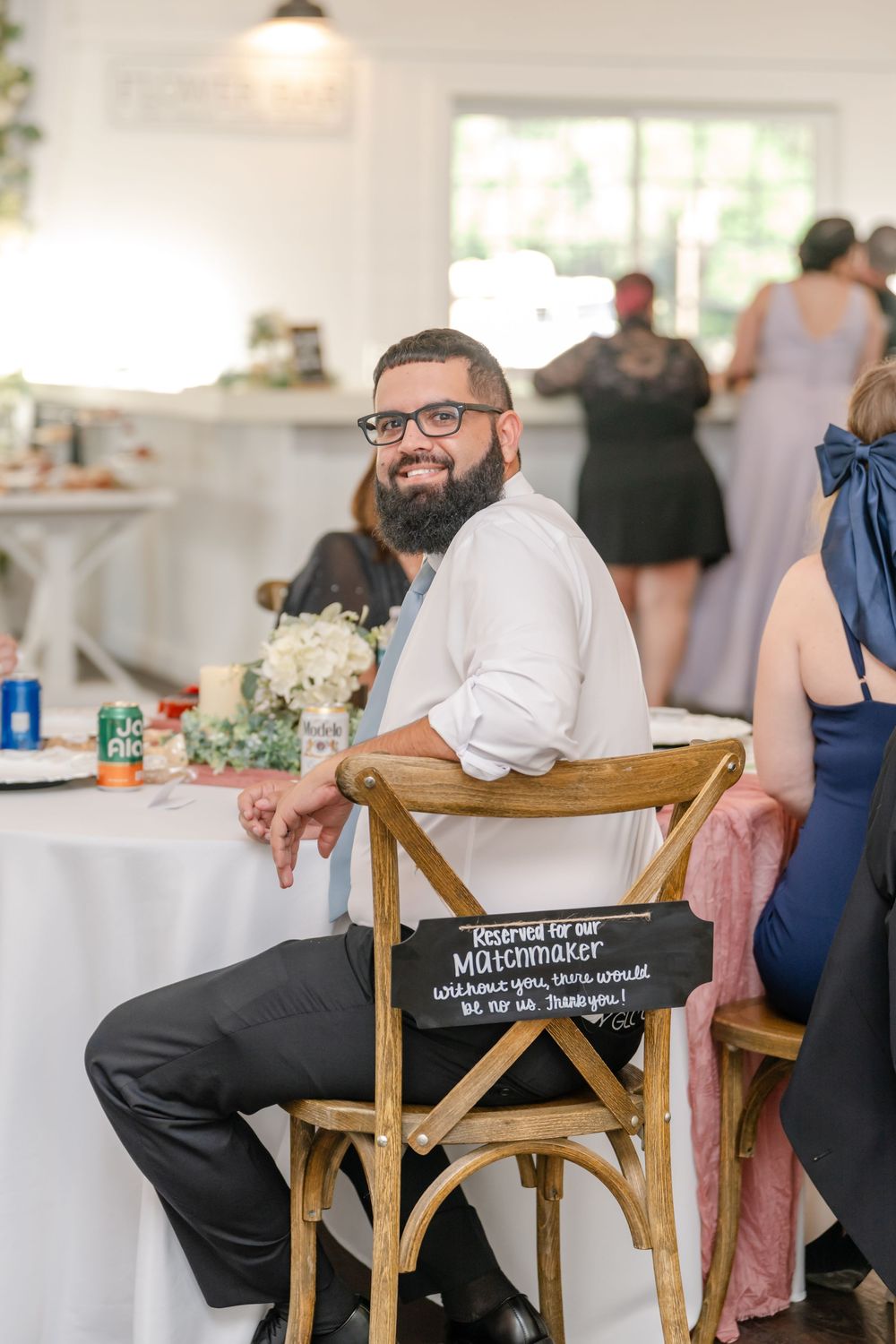 Bearded groom in white shirt smiles at camera while seated in wooden chair at wedding reception.